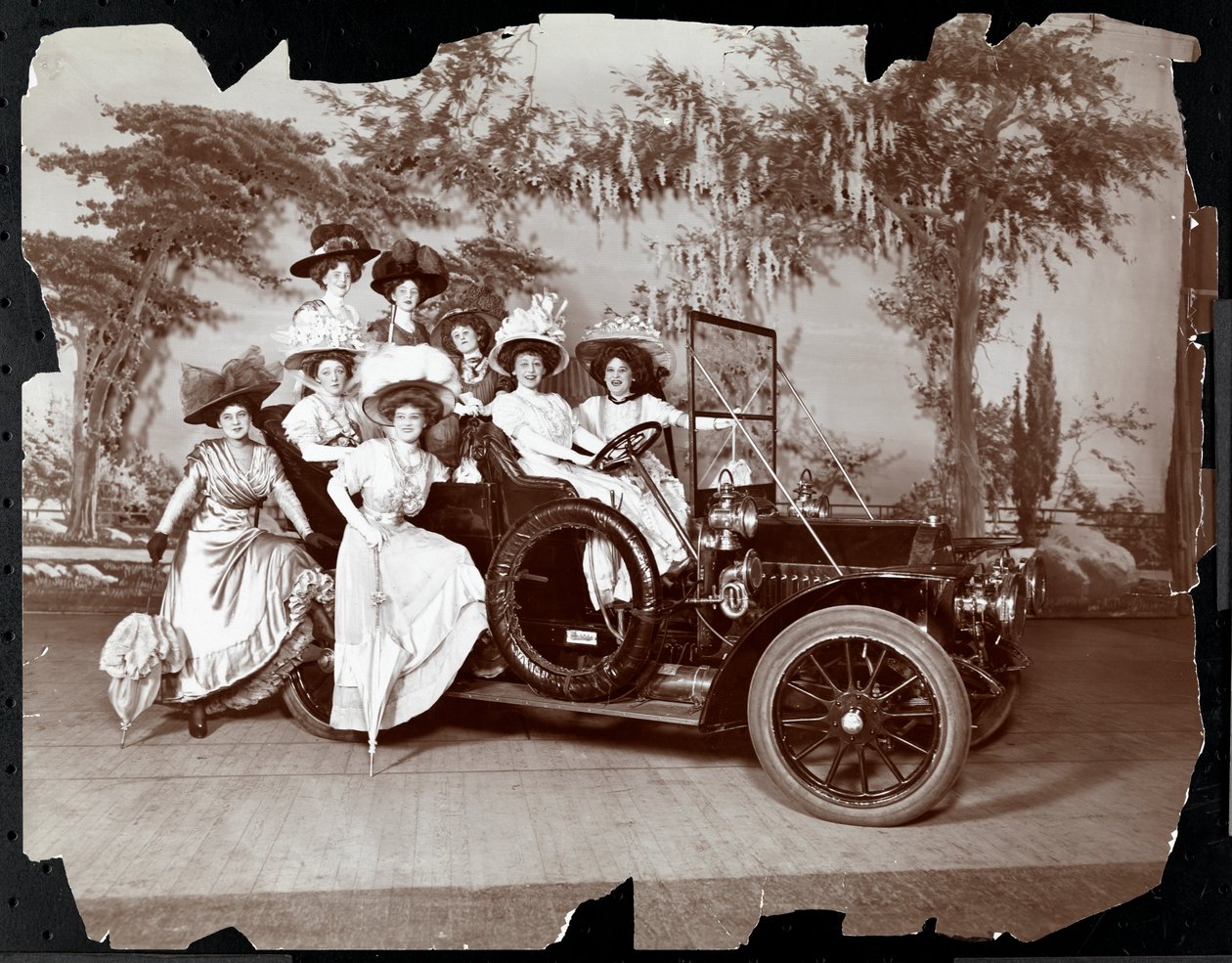 Osm žen v kostýmech pózuje v automobilu a kolem něj na jevišti s krajinnou kulisou, pravděpodobně při představení "Vanderbiltův pohár", 1906 (stříbrný želatinový tisk) od Byron Company
