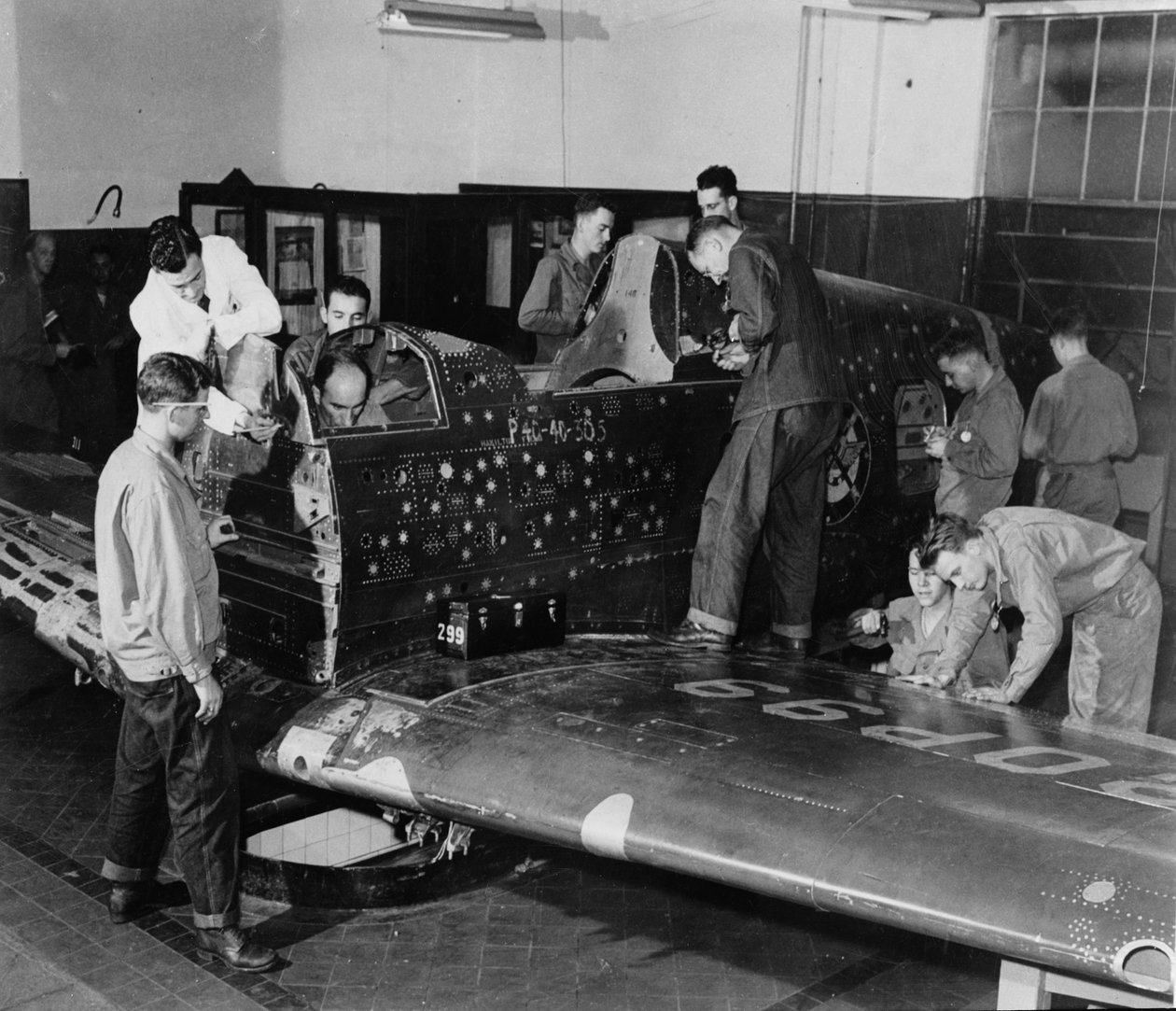 Vojenští stážisté pracují na „Flying Patch“, stíhacím letounu Curtiss „Warhawk“ bez motoru nebo ocasní části, se simulovanými dírami po kulkách a záplatami, kolem roku 1942 od American Photographer