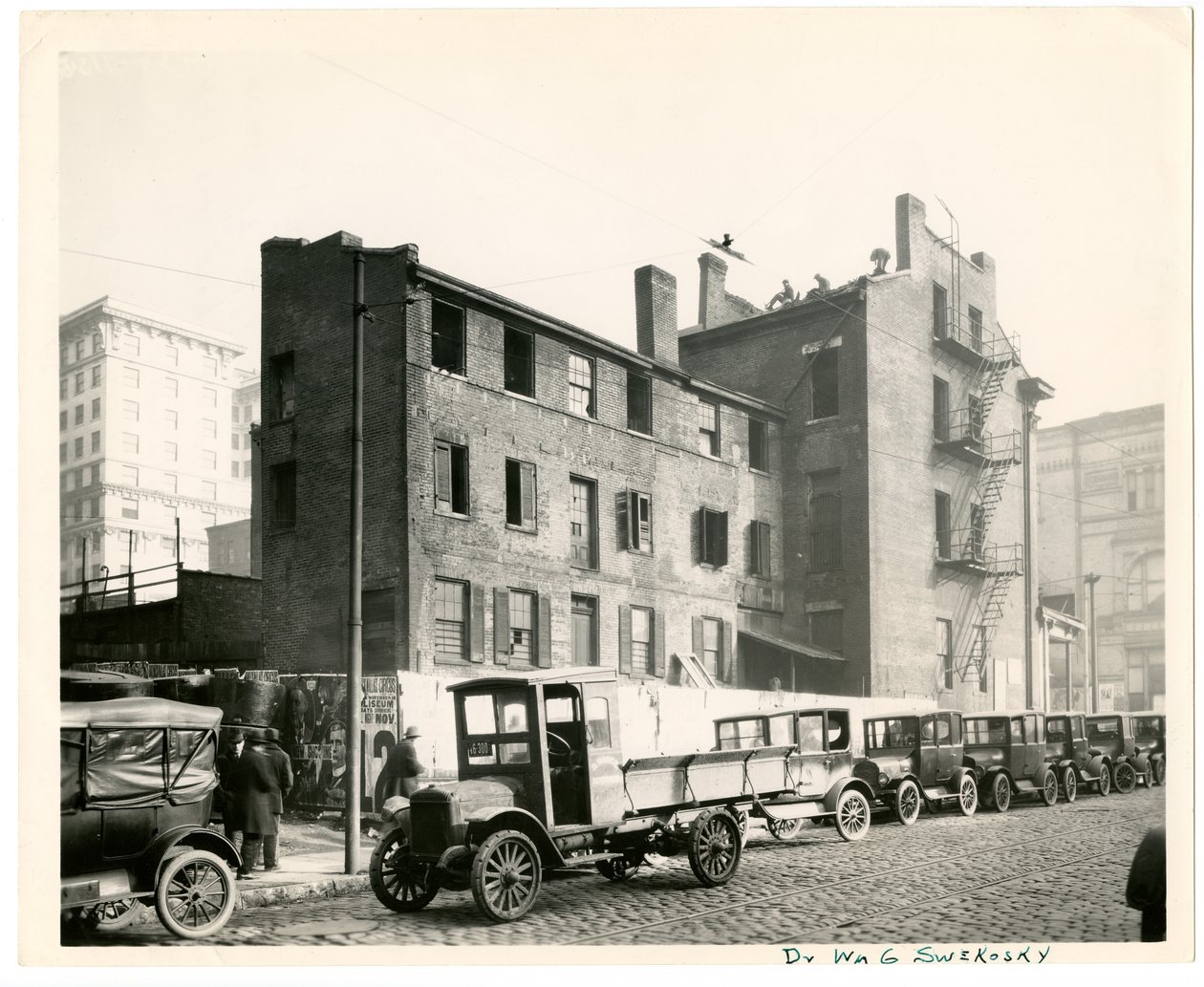 Horizontální, černobílá fotografie ukazující pohled zezadu na bar P. E. Burke v severozápadním rohu ulice 7th Street a Walnut od Charles Clement Holt
