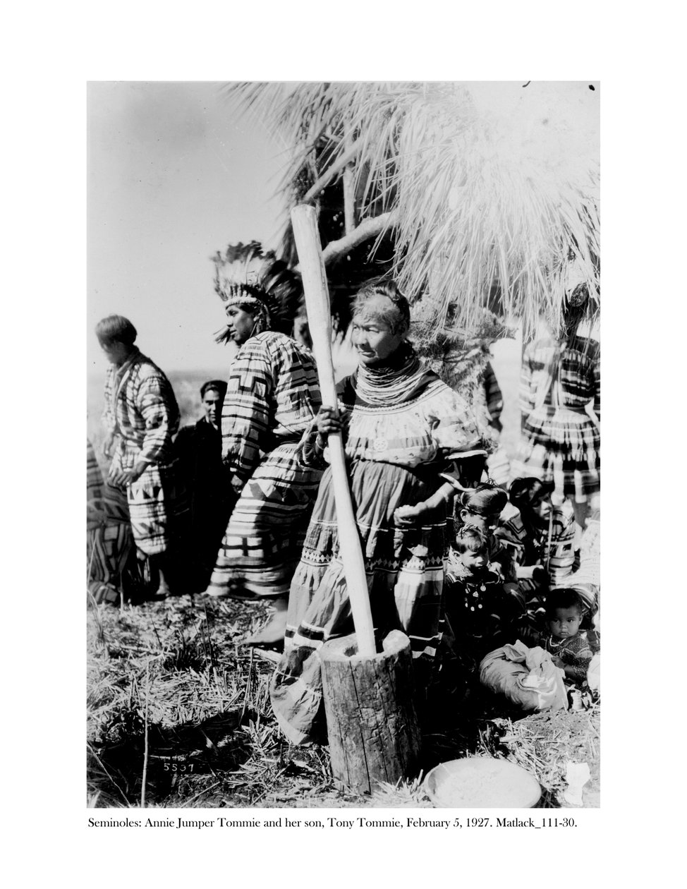 Annie Jumper Tommie pomocí hmoždíře a paličky k výrobě kukuřičné mouky a její syn Tony Tommie s čepicí z orlího peří, 5. února 1927 od Claude C. Matlack