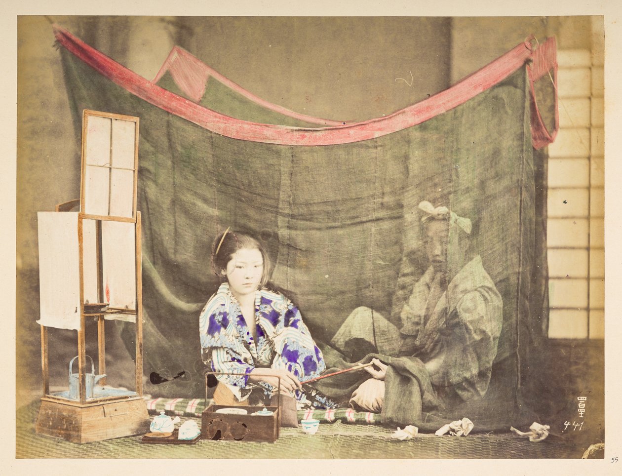 Japonský pár ve volném čase, Studiový obraz mladé Japonky, sedící s překážkou na přípravu čaje kolem sebe, podávající dýmku mužské postavě, která sedí za ní a je chráněna plátěnou moskytiérou,   Fotograf: Neznámý  Lucasova sbírka: od English Photographer