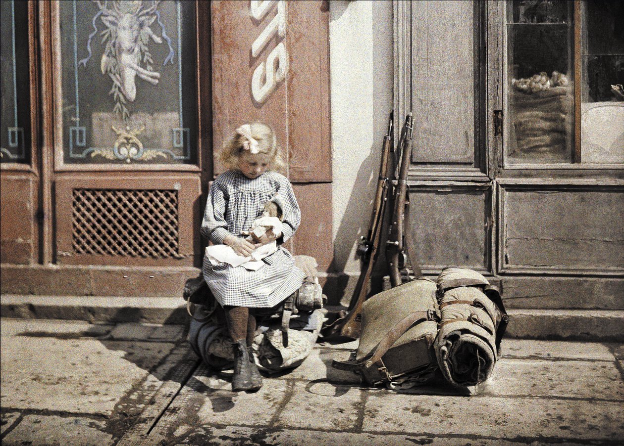 Holčička si hraje se svou panenkou; dvě zbraně a batoh jsou vedle ní na zemi, Reims, Marne, Francie, 1917 (autochrom) od Fernand Cuville