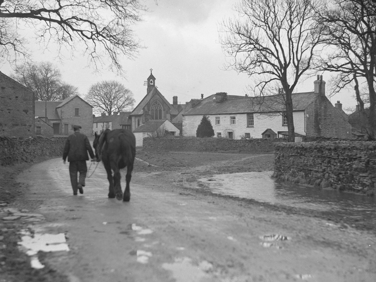 Muž vedoucí koně po silnici směrem od kamery k vesnickým stavbám a kostelu; architektura; náboženská; církevní; od Joseph Hardman