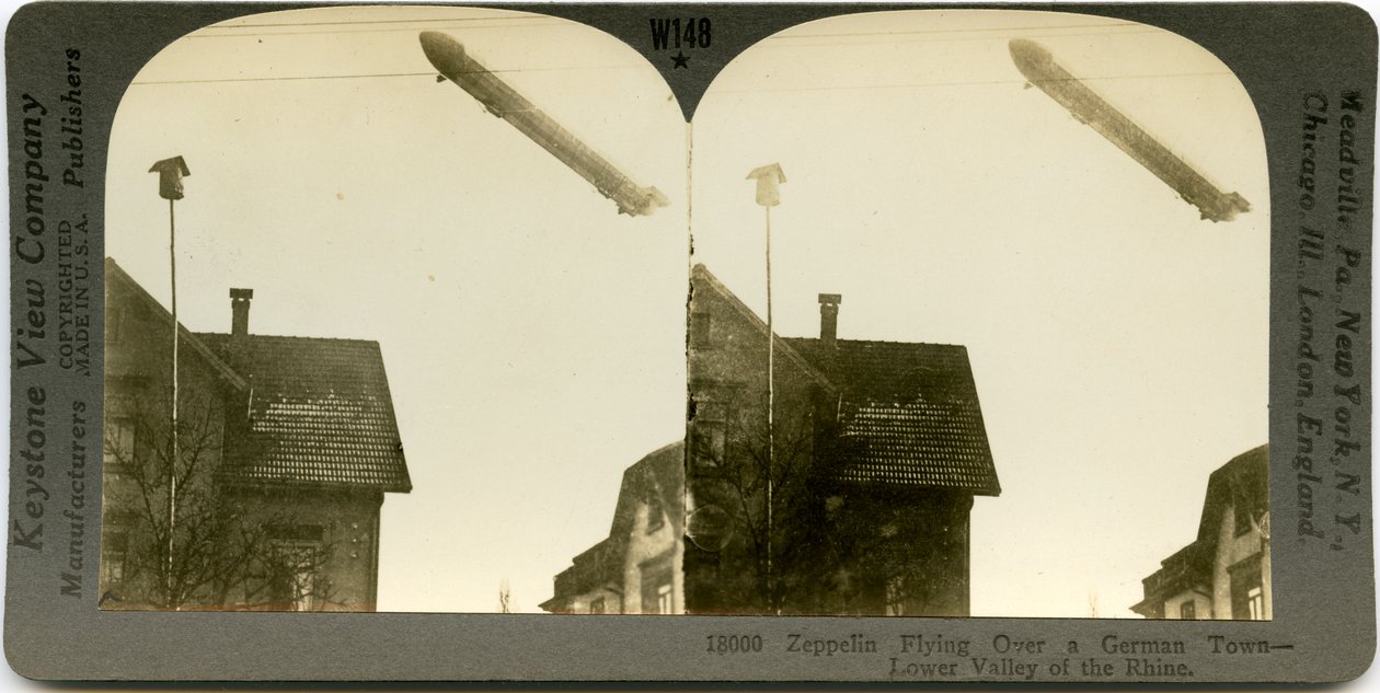 Horizontální, sépiová stereokarta ukazující let Zeppelina nad střechami města. Keystone Stereograf číslo 18000. Název zní od Keystone View Company