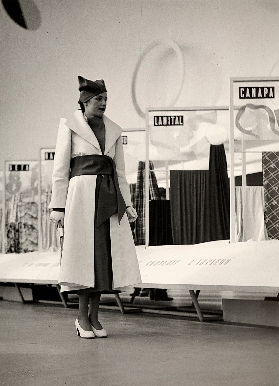Model v kabátě vytvořený couture house "Lamma". Výrobek je v pase nařasený širokou šnůrou a doplněn měkkým kloboukem. Fotografie byla pořízena v hale sloužící k výstavě látek. (černobílá fotografie) od Luis Bogino