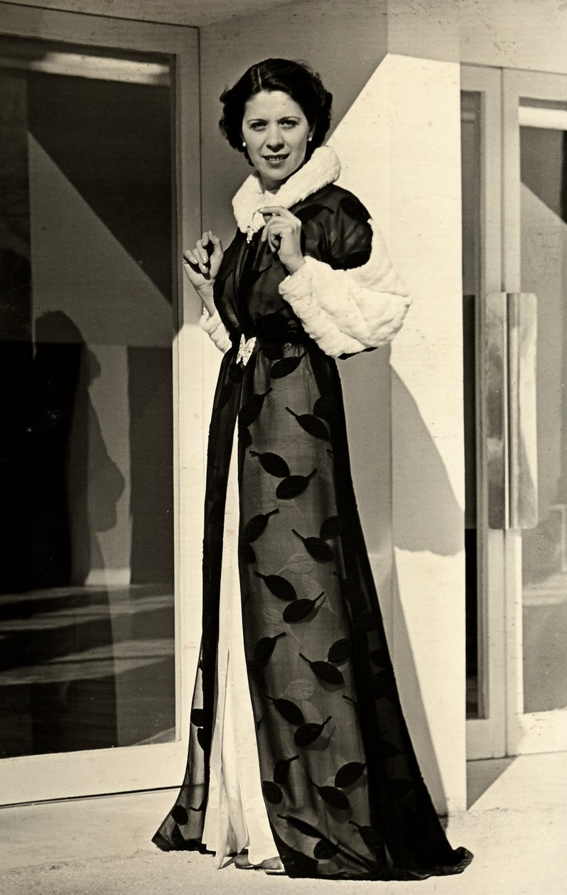 Modelka má na sobě večerní šaty vytvořené italským couture housem "Bernasconi". Prezentovaný výrobek se skládá ze šifonového kabátu s kožešinou lemovaným límcem a manžetami (č/b foto) od Luis Bogino