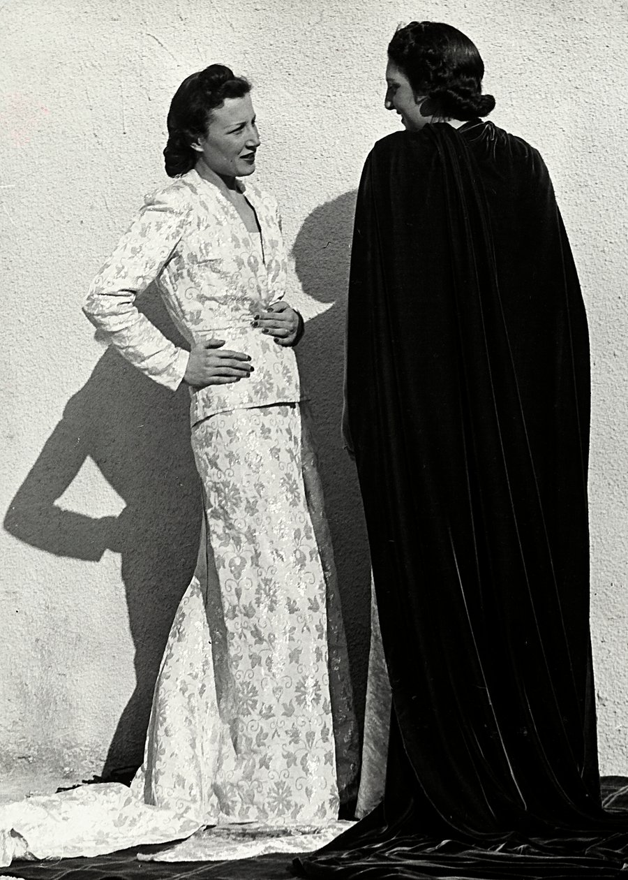 Dva modely představují výtvory italského módního domu "Dragoni". Jedna modelka má na sobě outfit z plenkového materiálu skládající se z bundy a dlouhé sukně s vlečkou. Druhý model, při pohledu zezadu, má na sobě dlouhý sametový plášť. (černobílá fotografie od Luis Bogino