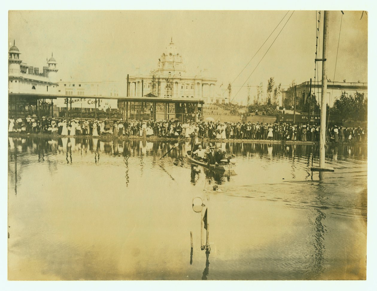 Horizontální fotografie plavce překračujícího cílovou čáru. Muži na lodi sledují stejně jako davy lidí na břehu od Unknown