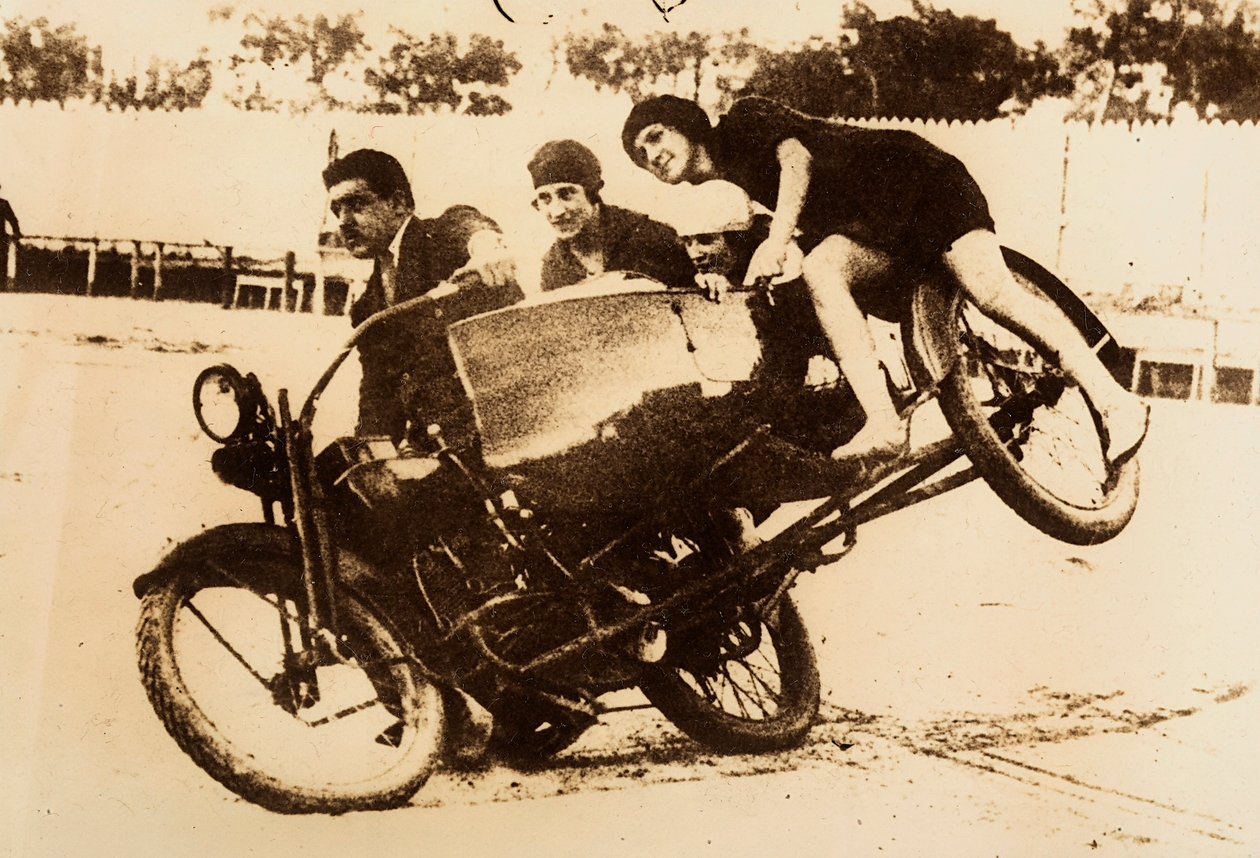 Muž předvádí jízdu na motocyklu s postranním vozíkem, na kterém jsou tři mladé ženy. Při zatáčení se sajdkára zvedá ze země. od Unknown photographer
