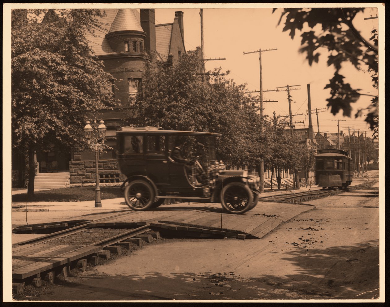 Horizontální, černobílá fotografie automobilu přecházejícího po kolejích trolejbusů, když se po trati pohybuje vozík od William G. Swekosky