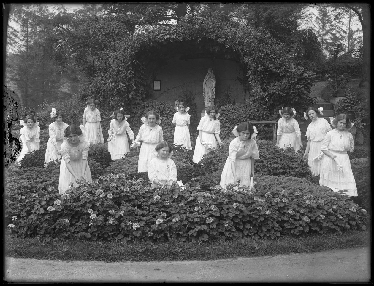 Maturitní ročník dívek z římskokatolického sirotčince v květinové zahradě před jeskyní Panny Marie, 1912 čb fotografie od William Davis Hassler