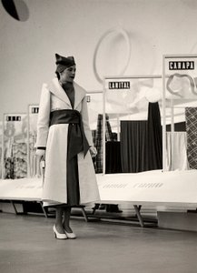 Model v kabátě vytvořený couture house "Lamma". Výrobek je v pase nařasený širokou šnůrou a doplněn měkkým kloboukem. Fotografie byla pořízena v hale sloužící k výstavě látek. (černobílá fotografie) od Luis Bogino
