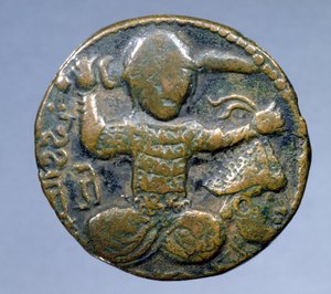 Líc mince (bronz) zobrazující Turka s přilbou sedícího se zkříženýma nohama, v pravé ruce drží meč za hlavou a v levé ruce useknutou hlavu v přilbě. razil Husam al-Din Yuluq Arslan (1184-1201)
