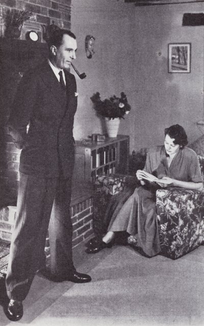 Manželka čte, zatímco manžel si zahřívá záda proti ohni, kolem roku 1950 od English Photographer
