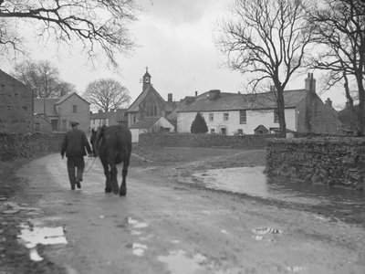 Muž vedoucí koně po silnici směrem od kamery k vesnickým stavbám a kostelu; architektura; náboženská; církevní; od Joseph Hardman