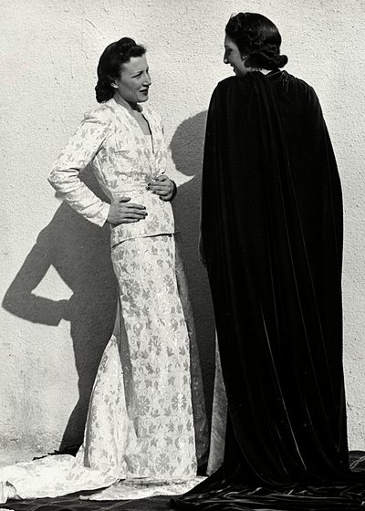 Dva modely představují výtvory italského módního domu "Dragoni". Jedna modelka má na sobě outfit z plenkového materiálu skládající se z bundy a dlouhé sukně s vlečkou. Druhý model, při pohledu zezadu, má na sobě dlouhý sametový plášť. (černobílá fotografie od Luis Bogino