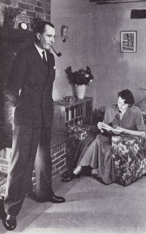 Manželka čte, zatímco manžel si zahřívá záda proti ohni, kolem roku 1950 od English Photographer