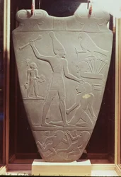 Narmerova paleta: ceremoniální paleta zobrazující krále Narmera s bílou korunou Horního Egypta, jak udeří na vězně, nalezená v Hierakonpolis, rané dynastické období, cca 3000 př. n. l. (pro podrobnosti)