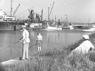 Muž a chlapec rybaří v Glasson Dock, zatímco přihlíží za nimi sedící dáma, několik lodí je zakotveno
