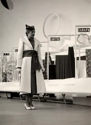 Model v kabátě vytvořený couture house "Lamma". Výrobek je v pase nařasený širokou šnůrou a doplněn měkkým kloboukem. Fotografie byla pořízena v hale sloužící k výstavě látek. (černobílá fotografie)