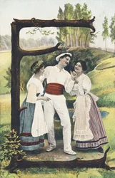 E: Muž oblečený v bílé a červené stojí mezi dvěma ženami v sukni, halence a zástěře. Venkovský dekor - abecední znaky Ref Th. E., L. Theochrom - Serie 994. Theodor Eisman, vydavatel, Lipsko