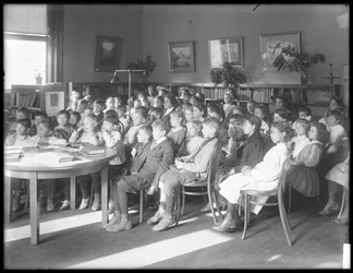 Děti sedí a poslouchají v čítárně neidentifikované pobočky veřejné knihovny Queens Borough, kolem roku 1910