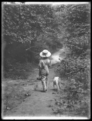 William Grey Hassler ve slaměném klobouku a kombinéze, nesoucí rybářský prut a košík, kráčí po lesní cestě s malým psem (Bessie), 20. srpna 1911