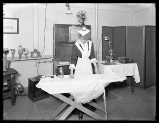 Mladá žena v kostýmu služebné předvádí použití elektrické žehličky, cca 1913-14